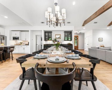 Bright open dining room with modern chandelier, exposed wood ceiling beams, twelve-seat farmhouse table, and kitchen and living areas visible in background at Yonah Mountain Farmhouse