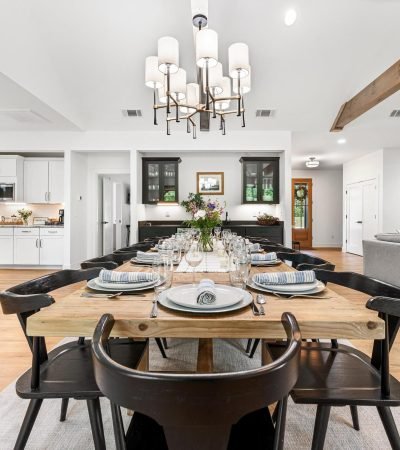 Bright open dining room with modern chandelier, exposed wood ceiling beams, twelve-seat farmhouse table, and kitchen and living areas visible in background at Yonah Mountain Farmhouse